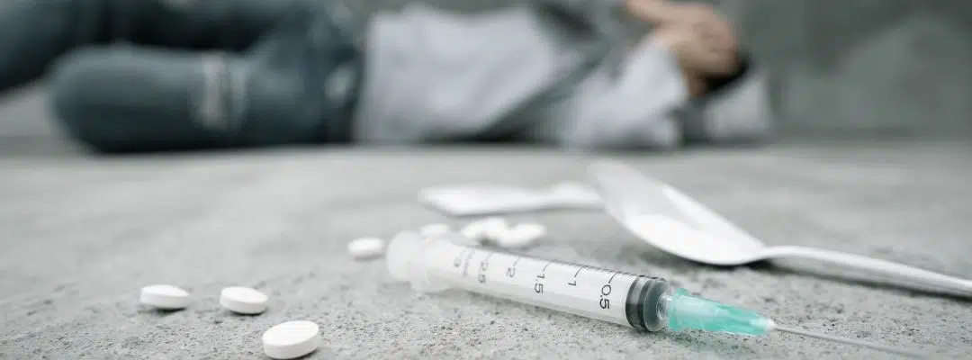 Image of a person laying in the background with a spoon, syringe and pills in the foreground - Hot Shots Are Most Dangerous for First-time Users