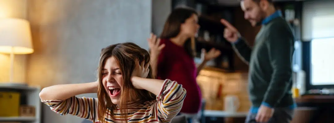 Image of a young girl screaming with her eyes closed and hands covering her ears, as her parents are seen arguing behind her
