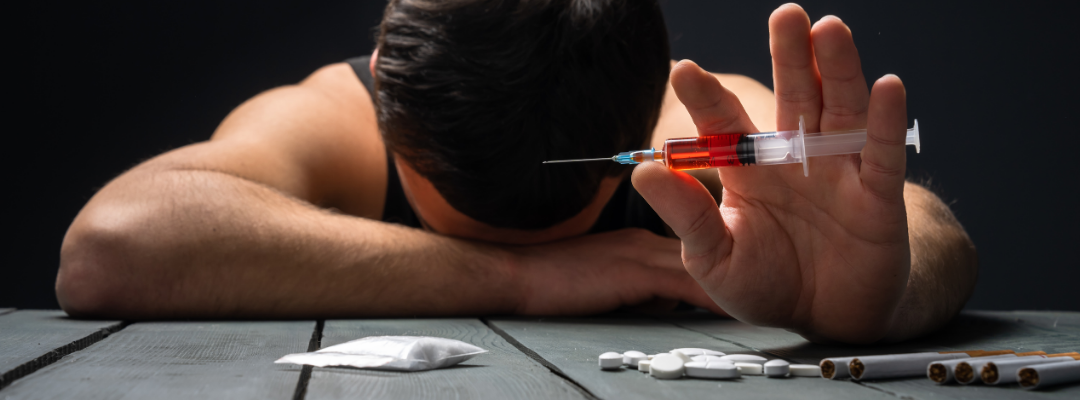 Image of a man holding a syringe with meth and white pills on the table in front of him, representing the dangers of combining opioids and stimulants