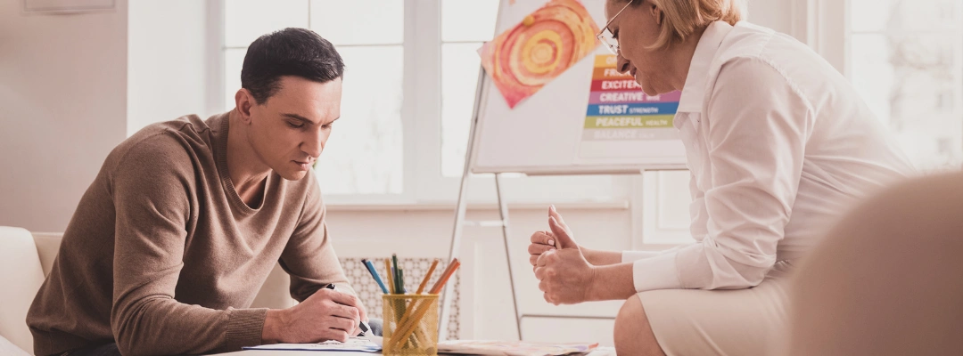 Image of a man in art therapy, drawing on a table as his therapist looks on