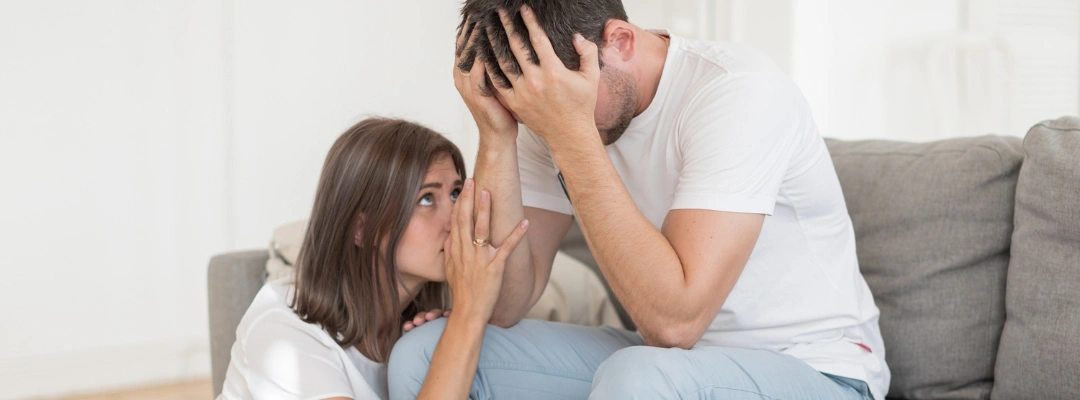 Image of a woman sitting on the floor beside a distraught man, who is sitting on a sofa with his hands covering his face